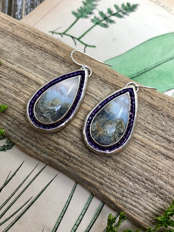 Two hand cut teardrops of scenic moss agate draw the eye in to their endless world of wonder. They are surrounded by petite amethyst gems and set in sterling silver.  Finished with sterling silver French wires. Perfect for everyday wear or a showstopping accessory for an evening on the town.

Product Details:

• Indonesian scenic moss agate
• 82 microfaceted amethyst gemstones
• Solid sterling silver
• French ear wires