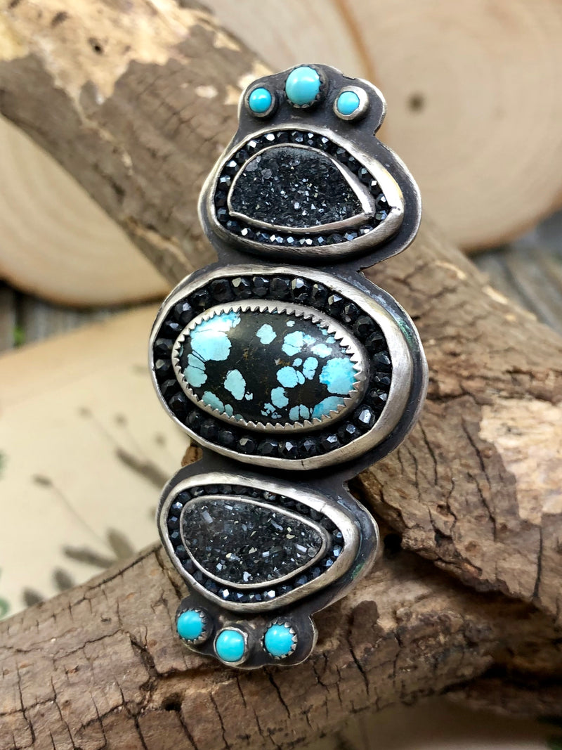When South Western-inspired jewelry has a dramatic flair, you are presented with the Western Gothic Ring. Blue pools of turquoise are surrounded by an expertly cut black spinel halo. This ring goes so many steps further with black agate druzy accent gems and hematite halos for added sparkle.  Finished with three sleeping beauty turquoise dots on either end.  All hand-forged in sterling silver.  Finished with a split shank size 8.  Antiqued to show the fine detail.

Product Details:

• 