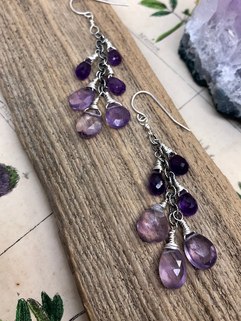 "Compassion is an action word with no boundaries" - Prince

Bask in the gentle glow of purple amethyst drops. Pear-shaped and faceted gems are wire wrapped along a silver chain and finished on standard French wires. A cascade of color perfect for any elegant ensemble. The perfect gift for a February birthday.

Product Details:

• Sterling silver ear wires
• Fourteen purple amethyst briolette gems
• Sterling and fine silver
• Approximately 2" long and just over 2.25" long from the top o
