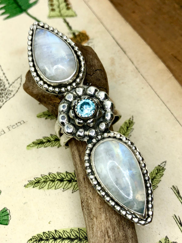Embrace an enchanted celebration of new life and divine awakening

Show off your inner goddess with this brilliant and bold over-size ring. Two iridescent rainbow moonstones embrace a glowing blue zircon. Each gem is skillfully set in a silver bezel and beaded surround. This size 8 ring is a perfect balance of shape and color, form and scale.

•Two drop-shaped rainbow moonstone gems
•One blue zircon
•Size 8 
•Fine and sterling silver