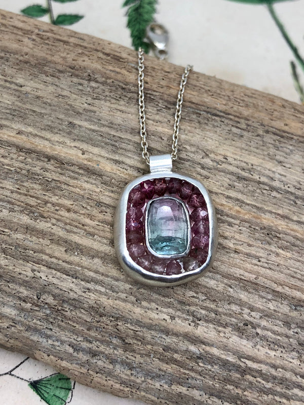 A rose cut bi-color tourmaline is surrounded by a halo of microfaceted ombré pink tourmalines and sterling and fine silver.

• Rose cut bi-color tourmaline 
• Shades of pink tourmaline gems
• Sterling and fine silver 
• Tube bail 

