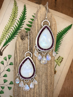 An elixir to smooth away any jagged edges.

Celebrate the glory and luminosity of the rising moon with teardrop rainbow moonstones. The subtle color scheme and gentle forms will soothe the senses as well as the soul. Theses earrings feature halos of contrasting rhodolite garnets and a constellation of 5 AAA rainbow moonstone briolettes on each earring.

• 2 Teardrop-shaped rainbow moonstones
• Faceted rhodolite garnet gems
• 10 AAA rainbow moonstone briolettes
• Fine and sterling silve